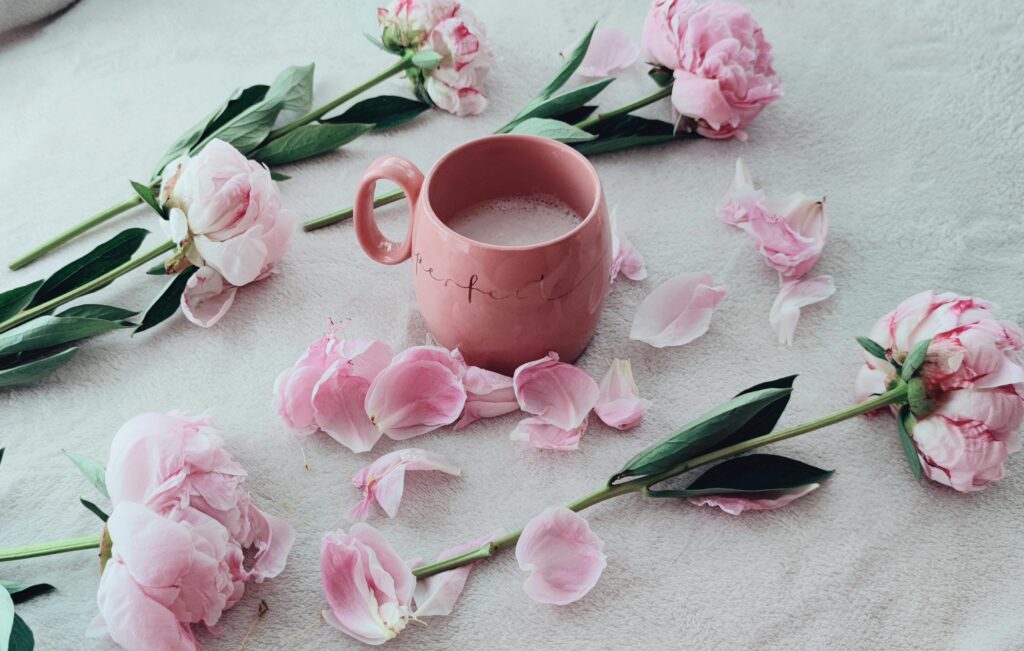 Aesthetic flat lay of pink mug surrounded by peonies and petals on fabric.